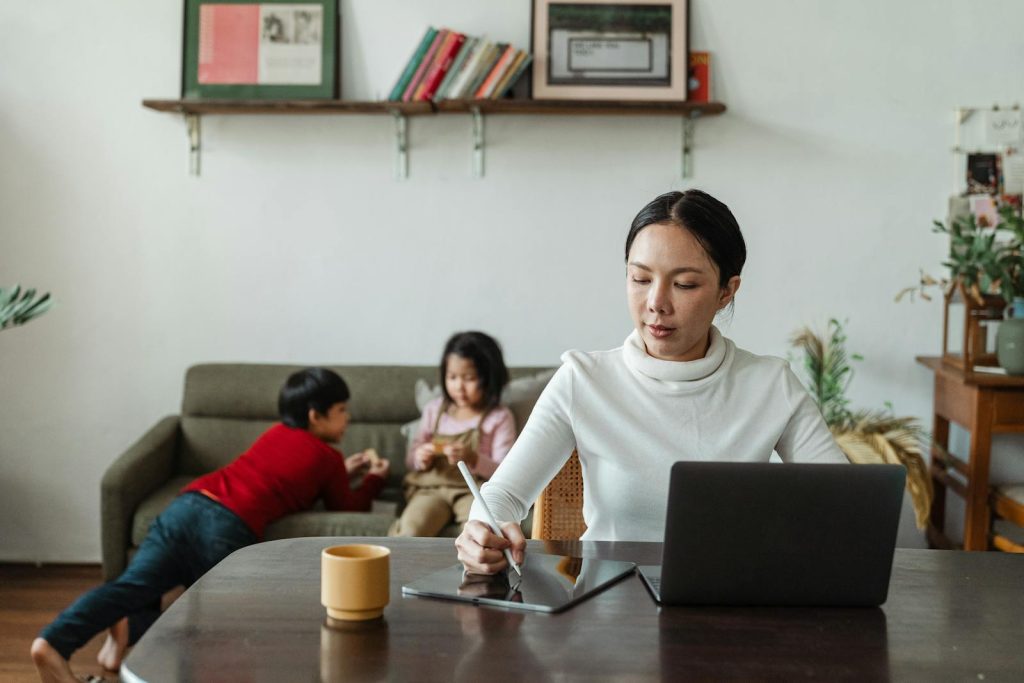 Mother multitasking with laptop and stylus alongside her children playing at home