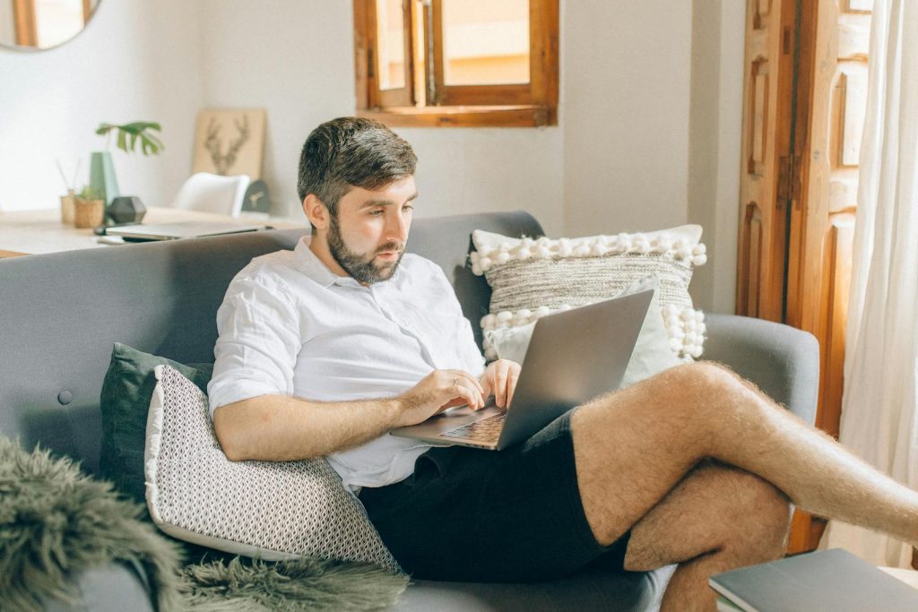 Adult man sitting on couch using laptop, working remotely from cosy home setting